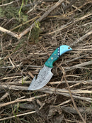 The Texan Knives Elite Custom Handmade Damascus Steel Guthook Skinning Knife, featuring a blue-green resin handle with white stars and a 7.5” premium blade, rests on dry twigs and grass outdoors.