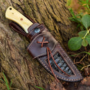The Texan Knives Premium Damascus Steel Skinner Knife features a custom handmade camel bone handle and comes sheathed in a brown leather case with decorative stitching and a black textured panel, displayed against a tree trunk with green leaves nearby.