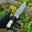 A gloved hand holds the Texan Knives Custom Handmade Skinning Knife with a 4.5" Damascus blade and a camel bone & resin handle in black, white, and red. A tree branch and green foliage appear in the background.