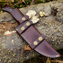 A brown leather sheath with brass buttons rests outdoors on a weathered tree trunk beside foliage and bones—an ideal match for your Texan Knives Custom Handmade 4.5-Inch Damascus Steel Skinning Knife with G10 Handle.