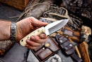 A hand wearing a silver watch holds the Texan Knives Custom Handmade J2 Steel Skinning Knife – 4.5" blade with an engraved camel bone handle. Nearby are outdoor gear items: a leather sheath, bag, and American flag patch.