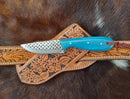 The Texan Knives Handcrafted Western Belt Knife with a blue handle and patterned blade rests on its floral-embossed brown leather sheath, making it an ideal year-round outdoor companion by Texan Knives.