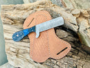 The Texan Knives Custom Handmade Bull Cutter Knife – Damascus Steel Holiday Edition features a blue and black handle, displayed on a stitched brown leather sheath atop textured wood.
