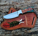The Texan Knives Rustic Cowboy Knife with a patterned multicolored handle and textured blade rests on its handcrafted, feather-painted leather sheath, both displayed on rugged tree bark for an authentic Western vibe.
