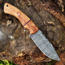The Texan Knives Custom Handmade 10" Damascus Steel Hunting Knife, featuring an olive wood handle and leather sheath, is displayed on a rugged tree trunk.