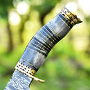 Close-up of the Texan Knives Vintage Damascus Steel Bowie Knife for Sale – 13", featuring a brass guard and a polished camel bone handle, set against a blurred green outdoor background.