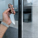 A hand wearing a silver watch holds the Texan Knives Handmade Cowboy Belt Knife with an 8" rasp steel blade and wooden handle, reflected in glass against a stone background. Comes with a Western leather sheath.