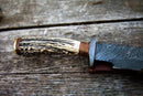 A close-up of the Texan Knives Damascus Steel 1800s Bowie Knife (15"), featuring a stag horn handle and a Damascus steel blade and guard, resting on weathered wood. Includes leather sheath.