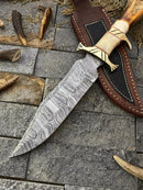 The Texan Knives Custom Handmade Damascus Steel Bowie Knife features a 13-inch patterned blade and a bone-and-brass guard handle. It rests on gray stone near antler pieces and comes with a brown leather sheath.