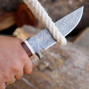 A hand uses the Texan Knives Damascus Steel Hunting Knife – 10" with patterned blade, Damascus steel guard, and stag horn handle to cut thick rope; blurred wooden logs form the background. Includes leather sheath.