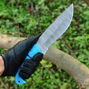 A hand in a black, textured glove holds the Texan Knives Custom Handmade Damascus Steel Hunting Knife – 10-Inch Blade with Resin Handle and Leather Sheath outdoors, with green foliage and tree branches in the background.