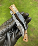 A person wearing a black glove holds a Texan Knives Custom Handmade Damascus Blade EDC Folding Pocket Knife with a color bone handle and brass engraved bolster, set against a blurred green background.