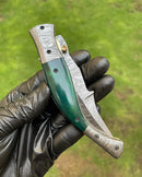 A gloved hand holds the Texan Knives Custom Handmade Damascus Blade EDC Folding Pocket Knife, featuring a 3-inch patterned blade, color bone handle, and brass engraved bolster, shown closed against a blurred green background.