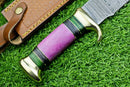 A close-up of the Texan Knives Damascus Steel "The Bowie Knife" with a 16" blade, brass guard, purple and green resin pakkawood handle, lying on grass beside its brown patterned leather sheath.