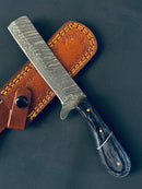The Texan Knives Handmade Damascus Steel Calf Cutter Knife features a 5" patterned blade and a dark Pakkawood handle, accented with brass pins, resting on a brown leather sheath with yellow stitching.