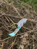 The Texan Knives Custom Handmade Damascus Steel Guthook Skinning Knife with a 7.5” resin handle and leather sheath rests on dry branches and leaves outdoors, showcasing its intricate blade pattern.