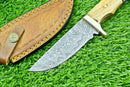 The Texan Knives Handmade Damascus Steel Hunting Knife features a patterned blade, an olive wood & resin handle, and comes with a brown leather sheath with yellow stitching, displayed on green grass.