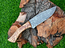 The Texan Knives Exquisite 10-Inch Damascus Steel Hunting Knife features a patterned blade, rosewood handle, and comes with a stylish leather sheath—an ideal outdoorsmen gift. Shown resting on tree bark in green grass.