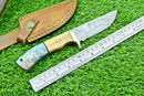 The Texan Knives Handmade Damascus Steel Hunting Knife features a patterned 23cm blade, an olive wood and resin handle, and comes with a leather sheath. A metal ruler lies nearby on green grass.