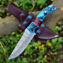 A Texan Knives Custom Handmade Damascus Steel Hunting Knife – 10-Inch Blade with a blue and red resin handle rests on a tree branch, secured in its brown leather sheath. Green foliage is softly blurred in the background.