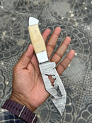 A hand holds the Texan Knives Custom Handmade Damascus Steel Knife in a Belt, featuring a patterned blade with cutout design and a light camel bone handle. The background shows a decorative, patterned surface.