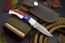 The Texan Knives Handcrafted Damascus Steel Pocket Knife – Texas Flag Handle & Leather Sheath rests on a wooden block with coiled rope and autumn leaves nearby, making it a standout collectible EDC gift.