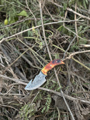 The Texan Knives Custom Handmade Damascus Steel Guthook Skinning Knife features a 7.5” blade, camel bone handle, and comes with a leather sheath, shown resting among dry branches and leaves outdoors.
