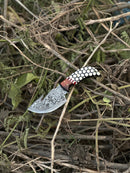 The Texan Knives Custom Handmade Damascus Steel Guthook Skinning Knife, featuring a 7.5" patterned blade and a rosewood & resin handle with leather sheath, is displayed outdoors on dry branches and leaves.