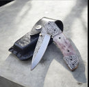 The Texan Knives Texas Knives Custom Handmade Engraved Pocket Knife features a D2 steel blade and engraved steel handle, shown partly open on a black leather sheath atop a light gray stone surface.