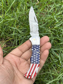 A hand holds the Texan Knives Lone Star 3-Inch Damascus Pocket Knife, custom handmade with a bone handle and open silver blade, against green grass.