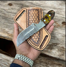 A hand holds the Texan Knives Handmade Cowboy Belt Knife, featuring a rasp steel blade and buffalo horn handle, above its leather sheath, all resting on a wooden surface.