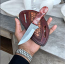A hand holds the Texan Knives Premium Handmade Western Knife—a Damascus blade with a resin & rosewood handle, resting on a tooled leather sheath. The person wears a silver wristwatch; a sink and countertop are in the background.