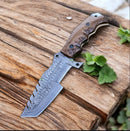 The Texan Knives Handmade Tom Brown Tracker Knife features a Damascus steel blade and rugged G10 handle, displayed on a rustic wooden surface beside green leaves to highlight the craftsmanship of this survival knife.
