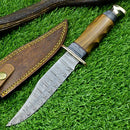 The Texan Knives Western Boulder Colorado Knife features an 11" handmade Damascus steel blade, olive wood handle, and brown leather sheath with yellow stitching, showcasing superior craftsmanship on a backdrop of green grass.