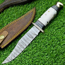The Texan Knives Hand-Forged Western Boulder Colorado Knife features an 11" Damascus steel blade, a resin grip with white, black, and brown accents, and a brass guard. It rests next to its brown leather sheath on green grass.