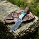The Texan Knives Handmade Western Damascus Knife features a decorative patterned blade and a resin and rosewood handle, displayed on its leather sheath atop a rough stone outdoors—a striking blend of artistry and rugged elegance from Texan Knives.