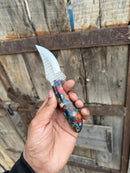 A hand holds the Texan Knives Premium Custom Handmade Western Cowboy Knife, featuring a durable rasp tool blade and colorful resin handle, in front of a weathered wooden gate.