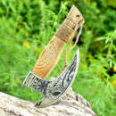 The Texan Knives Custom Hand Engraved Ax Pizza Cutter features a carved wooden handle and intricate engravings on its J2 steel blade, beautifully displayed resting on a log with green foliage in the background.
