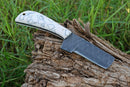 The Texan Knives Custom Handmade Cowboy Bull Cutter Knife features a 4" black textured J2 steel blade and a white stone-inlay handle, displayed on a weathered log with green grass in the background.