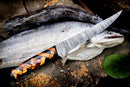 A fresh fish lies on wood with the 13" Custom Handmade Damascus Steel Fillet Knife by Texan Knives, showcasing its geometric handle. Sawdust and a green leaf nearby highlight the unique artistry of this premium fillet knife for sale.