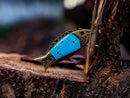 The Texan Knives Custom Handmade Fish Style Damascus Steel Folding Knife features an epoxy resin handle with brass accents and comes with a leather sheath—its striking design makes it stand out in any outdoor setting.