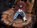 The Texan Knives Custom Handmade Fish Style Damascus Steel Folding Knife with a sheep horn & brass handle rests on a tree stump, its leather sheath upright behind it.