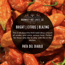 Close-up of vivid orange chili peppers and a circular black label with text about Harmacy Hot Sauce Co. Pata del Diablo Extra Hot Tomatillo & Clementine Sauce 9oz—a zesty, smoky, citrusy sauce perfect for heat lovers.