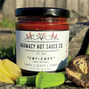 A jar of Harmacy Hot Sauce Co. Chi-Chee Kimchi Sauce 9oz is placed on stone outdoors, surrounded by fresh ginger, green onion, and a shiitake mushroom, showcasing this bold fermented hot sauce in the sunlight.