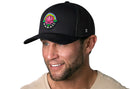 A man with a trimmed beard smiles while wearing the Haka Hat Hawaii Aloha Trucker Hat—black, featuring a colorful HI flower and "Hawaii" embroidery. He pairs it with a beige shirt against a white background.