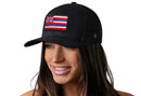 A woman with long brown hair smiles while wearing the Haka Hat Hawaii Flag Trucker Hat, a black HI snapback by Haka Hat featuring the Hawaiian flag patch on the front. The background is white.