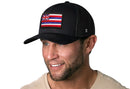 A man with light facial hair wears a Black HI Snapback Haka Hat Hawaii Flag Trucker Hat by Haka Hat, smiling in a beige shirt against a white background.