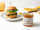 A jar of Terrapin Ridge Farms Amber Ale Pineapple Jalapeño Jam with a knife topped with jam sits beside a burger with lettuce, red onion, and cheese—ideal for adding sweet heat or as a flavorful grilling glaze.