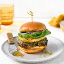 A gourmet burger with a brioche bun, grilled patty, spinach, Terrapin Ridge Farms Amber Ale Pineapple Jalapeño Jam, melted cheese, and sauce is skewered on a plate with knife and relish; drinks and a cloth appear in the background.