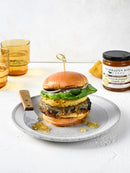 A burger with lettuce, grilled pineapple, and sweet-heat sauce sits on a bun atop a plate with a knife. Two yellow glasses and Terrapin Ridge Farms Amber Ale Pineapple Jalapeño Jam are in the background, some jam spilled on the plate.